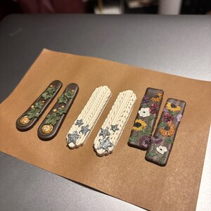 Puede incluir: Conjunto de seis pendientes hechos a mano de arcilla polimérica. Dos pares presentan un fondo marrón con hojas verdes y una calabaza dorada. Un par presenta un fondo blanco con un patrón de punto de jersey y estrellas plateadas brillantes. El último par presenta un fondo marrón con un diseño floral en tonos de rosa, naranja y amarillo.