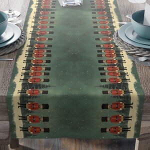 May include: A green table runner featuring a repeating pattern of toy soldiers in red coats and black hats. The runner is set on a wooden table, with place settings and a vase of pink flowers.