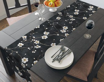 May include: A black table runner with a white floral pattern, placed on a dark wooden table. The runner features delicate white blossoms and branches. The table is set with plates, silverware, wine glasses, and a bowl of fruit.
