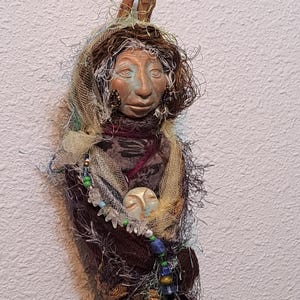 May include: A handcrafted doll with a serene face, adorned with a fabric scarf, beaded necklace, and earrings. The doll's head is topped with a twig and surrounded by wispy fibers. The color palette includes earthy tones, blues, and greens, creating a whimsical, artistic piece.