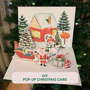 Puede incluir: Una tarjeta de Navidad pop-up DIY con una casa roja con nieve, un muñeco de nieve, Papá Noel, un trineo y regalos. La tarjeta tiene un diseño festivo con árboles de Navidad y un letrero. El texto "DIY POP-UP CHRISTMAS CARD" está en la parte inferior.