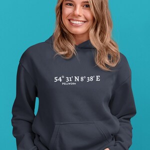 Könnte beinhalten: Ein marineblauer Kapuzenpullover mit einer Vordertasche. Der weiße Text auf der Vorderseite lautet "54° 31' N 8° 38' E" über "PELLWORM". Der Hintergrund ist türkis.