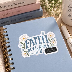 Peut inclure: Un carnet à spirales bleu clair orné d'un autocollant avec le texte Faith over fear, des illustrations florales et une petite étiquette noire indiquant The Church of Jesus Christ of Latter-day Saints, posé près d'un stylo blanc et de livres.