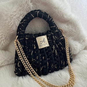May include: Black handbag with gold accents and a gold chain strap. The bag has a textured, woven appearance and a short handle. A small tag reads "A Authentic by JM".