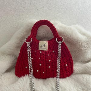 May include: A red crocheted handbag with a rounded top handle and silver chain strap. The bag is embellished with small white pearls and a tag that reads "Authentic by Dm". The bag is sitting on a white, fluffy surface.