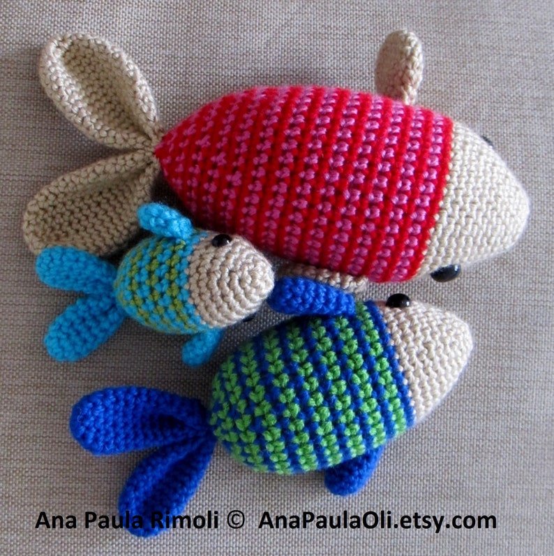 May include: Three crocheted fish toys. The largest fish is red and pink with beige fins. The medium fish is beige with blue and green stripes and blue fins. The smallest fish is beige with green and blue stripes and blue fins.