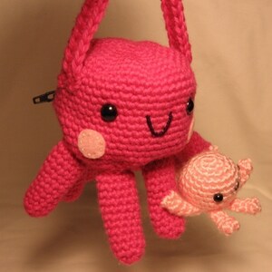 Puede incluir: Un pulpo de crochet rosa con una cara sonriente y un pulpo de crochet rosa m&aacute;s peque&ntilde;o unido a uno de sus brazos. El pulpo m&aacute;s grande tiene una cremallera en la espalda y una correa para llevar.