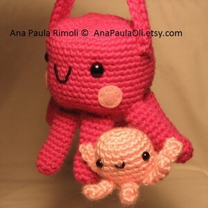 Puede incluir: Un pulpo de crochet rosa con un pulpo de crochet rosa m&aacute;s peque&ntilde;o unido a su brazo. El pulpo m&aacute;s grande tiene ojos negros y una nariz rosa. El pulpo m&aacute;s peque&ntilde;o tiene ojos negros y una boca rosa.