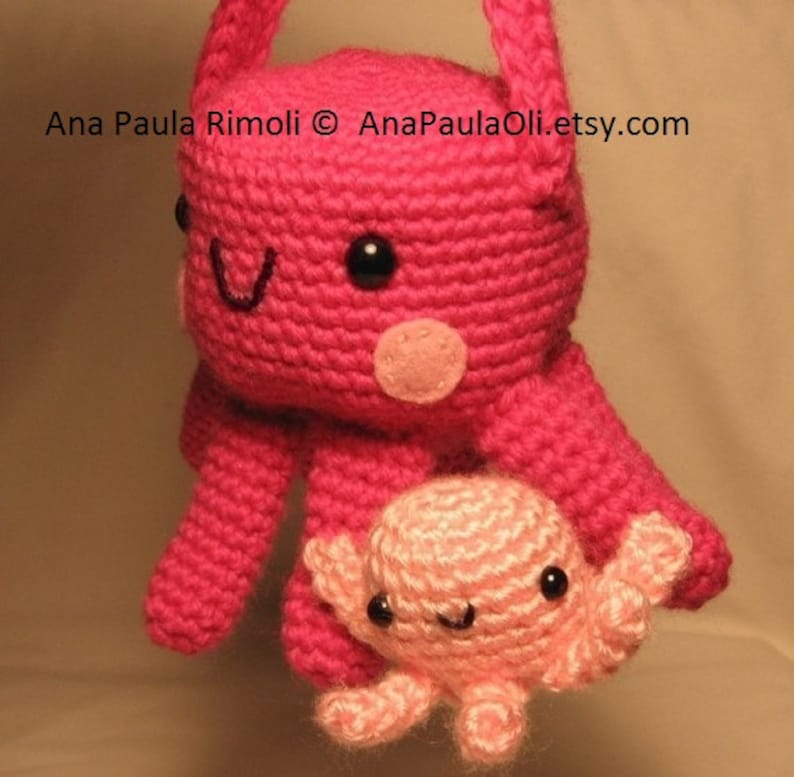 Puede incluir: Un pulpo de crochet rosa con un pulpo de crochet rosa m&aacute;s peque&ntilde;o unido a su brazo. El pulpo m&aacute;s grande tiene ojos negros y una nariz rosa. El pulpo m&aacute;s peque&ntilde;o tiene ojos negros y una boca rosa.