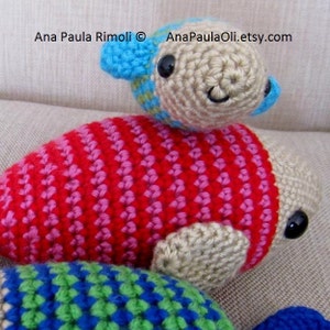 May include: Three crocheted fish toys. The largest fish is red and pink with white stripes. The middle fish is beige with blue and green accents. The smallest fish is beige with blue accents. The fish are on a beige fabric background.