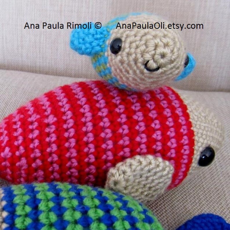 May include: Three crocheted fish toys. The largest fish is red and pink with white stripes. The middle fish is beige with blue and green accents. The smallest fish is beige with blue accents. The fish are on a beige fabric background.