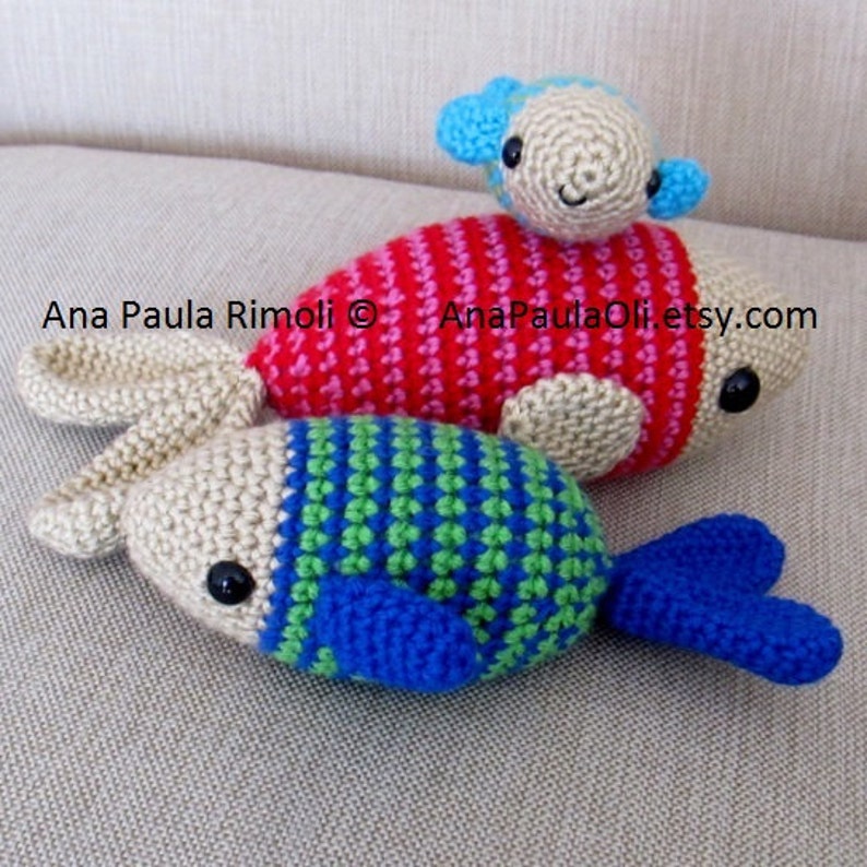 May include: Three crocheted fish toys. The largest fish is red and pink with a smaller beige fish on top. The smallest fish is beige with blue, green, and white stripes.