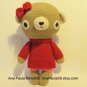 Puede incluir: Un oso de crochet marrón que lleva un vestido rojo y un lazo rosa. El oso tiene ojos negros y una nariz blanca.