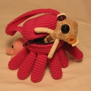 Puede incluir: Un pulpo de crochet rosa con una bolsa con cremallera. Un peque&ntilde;o pulpo de crochet rosa y un juguete de peluche beige con ojos negros y una nariz de bot&oacute;n amarillo est&aacute;n dentro de la bolsa.