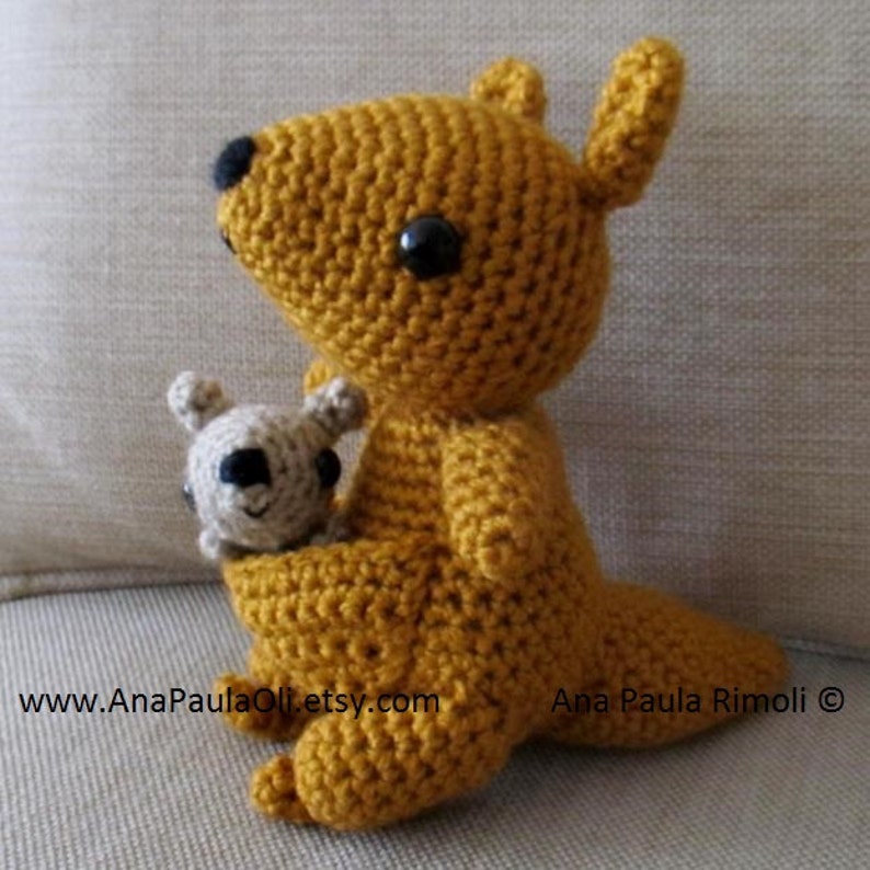 May include: A handmade crochet kangaroo in a mustard yellow color, with a smaller joey in its pouch. The kangaroo has black eyes and a black nose. The joey is a light beige color with black eyes. The Etsy shop URL is visible.