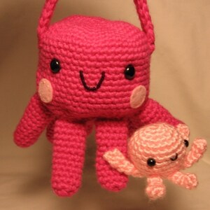 Puede incluir: Un pulpo de crochet rosa con un pulpo de crochet rosa m&aacute;s peque&ntilde;o unido a uno de sus brazos. Ambos pulpos tienen ojos negros y una boca sonriente.