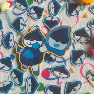 Könnte beinhalten: Eine Sammlung von Aufklebern mit einer Cartoonfigur in einem dunkelblauen Outfit mit einem Pik-Symbol. Die Aufkleber sind von anderen herzförmigen Aufklebern mit ähnlichem Design umgeben, alle auf einem gemusterten Hintergrund. Die Figur hat eine blaue Zunge und ein lächelndes Gesicht.