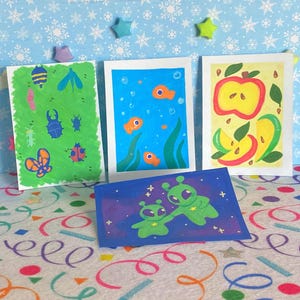 May include: Four colorful art prints featuring whimsical illustrations. One print showcases insects on a green background, another depicts fish in blue water, a third displays an apple and slices, and the last shows two green aliens on a blue background.