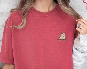 May include: A red-orange t-shirt with a small graphic of a cat on a skateboard. The shirt is short-sleeved and has a crew neck. The word "CRIMSON" is printed in red on the upper right corner.