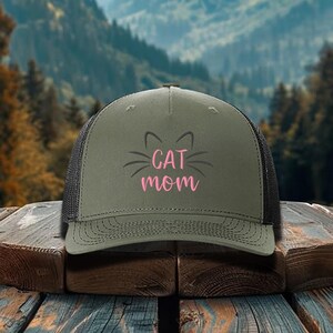 May include: A green and black trucker hat with a pink and black cat face and the text "CAT MOM" embroidered on the front.