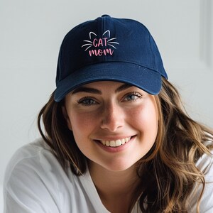 May include: A navy blue baseball cap with a white cat face and the text "CAT MOM" in pink lettering.