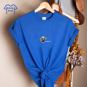 Puede incluir: Camiseta azul real con un peque&ntilde;o gr&aacute;fico de un vaso de cerveza y el texto "Vitamin G". La camiseta est&aacute; anudada a la cintura y cuelga de una percha de madera. Las palabras "Royal Blue" est&aacute;n en un cuadro azul en la esquina superior izquierda.