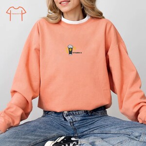 May include: Coral-colored sweatshirt with a white crew neck collar. The sweatshirt features a small graphic of a beer glass with the text "VITAMIN G" printed in black. The model is wearing blue jeans.