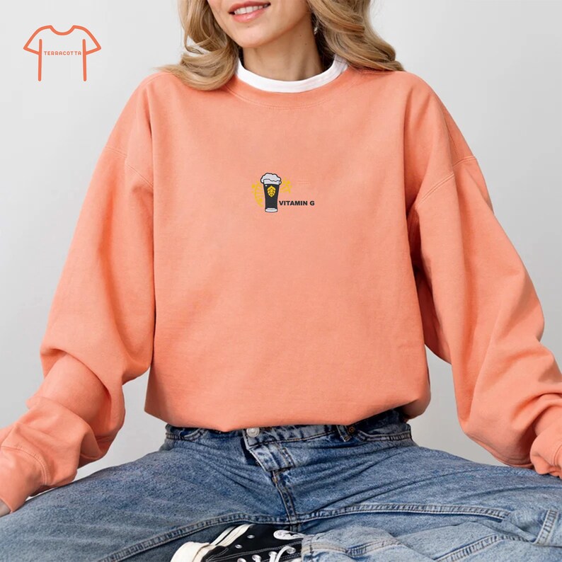 May include: Coral-colored sweatshirt with a white crew neck collar. The sweatshirt features a small graphic of a beer glass with the text "VITAMIN G" printed in black. The model is wearing blue jeans.