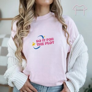 May include: A light pink t-shirt with the phrase "DO IT FOR THE PLOT" in pink text, a yellow star, and a blue crescent moon design. The shirt is part of a clothing collection.