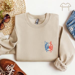 May include: A beige sweatshirt with a blue and red graphic design on the front. The graphic is a circle with a stylised design inside. The sweatshirt is made of a soft, comfortable material.