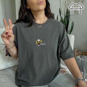 May include: Dark gray t-shirt with a small graphic of a beer glass with yellow accents and the text "VITAMIN G". The shirt is a comfortable, oversized fit.