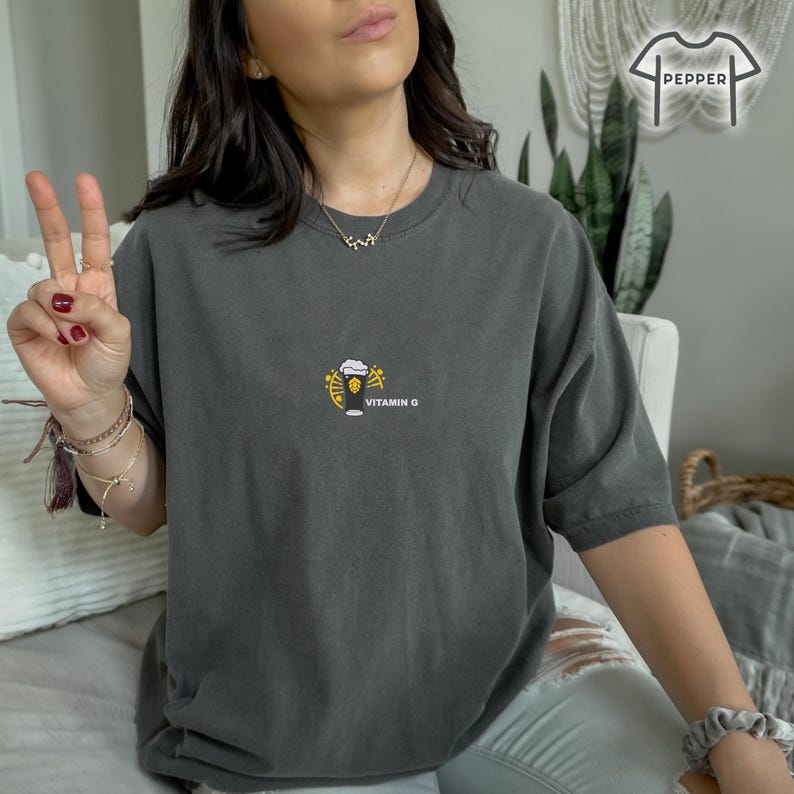 May include: Dark gray t-shirt with a small graphic of a beer glass with yellow accents and the text "VITAMIN G". The shirt is a comfortable, oversized fit.
