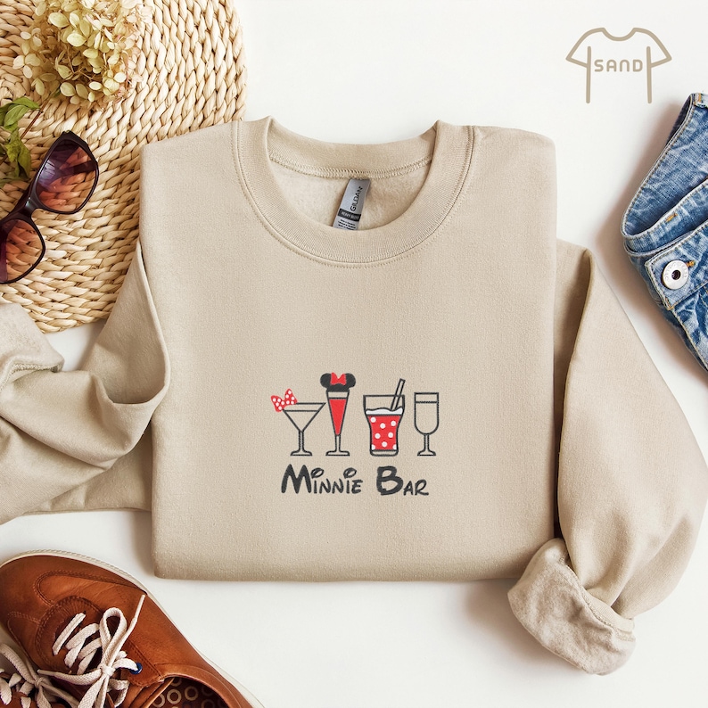 Puede incluir: Una sudadera beige con un gr&aacute;fico de Minnie Mouse y cuatro c&oacute;cteles. El texto "Minnie Bar" est&aacute; impreso debajo del gr&aacute;fico.