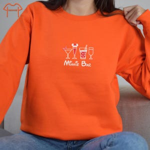 Puede incluir: Sudadera naranja con un gr&aacute;fico blanco de una fila de bebidas con el texto "Minnie Bar" debajo.