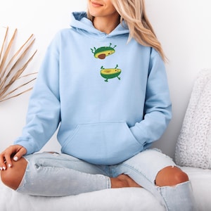 May include: A light blue hoodie featuring a playful avocado design. The design includes two cartoon avocados, one with a pit, and the other without. The hoodie has a kangaroo pocket and a drawstring hood. The model is wearing light blue jeans.