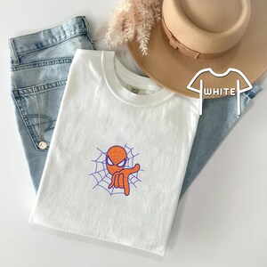 May include: White t-shirt with a cartoon image of Spiderman's head in a web. The image is in orange and purple.