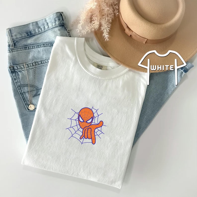 May include: White t-shirt with a cartoon image of Spiderman's head in a web. The image is in orange and purple.