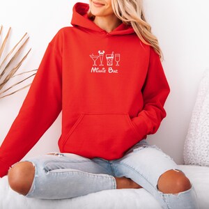 May include: A red hoodie with a front pocket and a drawstring hood. The hoodie features a white graphic of cocktail glasses and the text "Minnie Bar". The person is wearing ripped blue jeans.