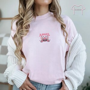 May include: Light pink t-shirt with the word "ANGEL" above a cartoon character. The character is pink with large ears and a small smile. The shirt is a soft, casual style.