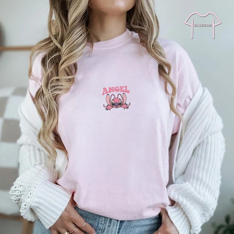 May include: Light pink t-shirt with the word "ANGEL" above a cartoon character. The character is pink with large ears and a small smile. The shirt is a soft, casual style.
