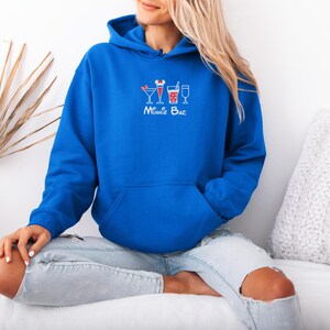 May include: A royal blue hoodie with a front pocket and a drawstring hood. The hoodie features a white graphic of three cocktail glasses with red accents and the text "Minnie Bar" in white script.