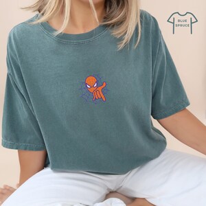 May include: A teal blue t-shirt with a cartoon image of Spiderman's head and hand in a web. The text "BLUE SPRUCE" is in the upper right corner of the image.