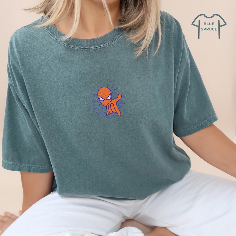 May include: A teal blue t-shirt with a cartoon image of Spiderman's head and hand in a web. The text "BLUE SPRUCE" is in the upper right corner of the image.
