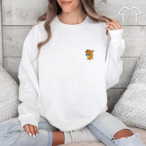 May include: White sweatshirt featuring a small cartoon graphic of an orange cat holding a blue book. The sweatshirt has a crew neck and long sleeves. The word "WHITE" is printed on the upper right corner.