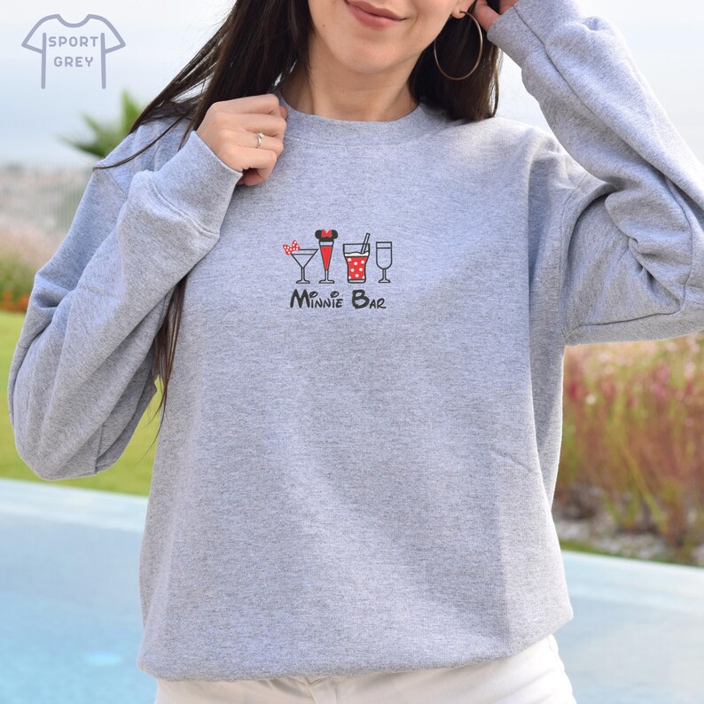 Puede incluir: Una sudadera gris con un dise&ntilde;o de Minnie Bar que presenta una cabeza de Minnie Mouse con lunares rojos y blancos con un lazo rojo y cinco iconos de bebidas diferentes. El texto "Minnie Bar" est&aacute; debajo de los iconos.