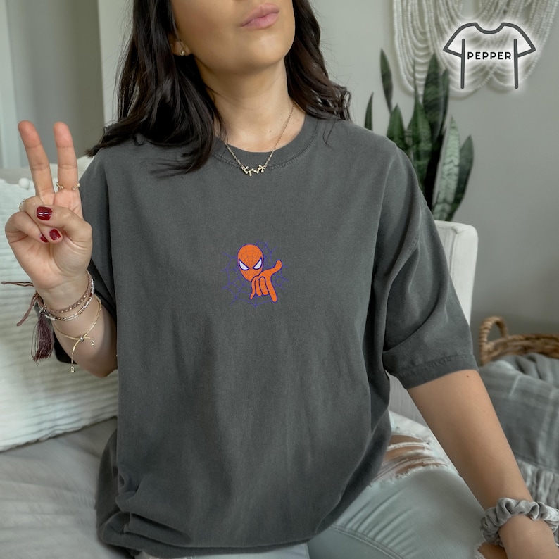 May include: A dark gray t-shirt with a small orange and purple graphic of a cartoon superhero character making a hand gesture. The character has a red mask and a white eye.