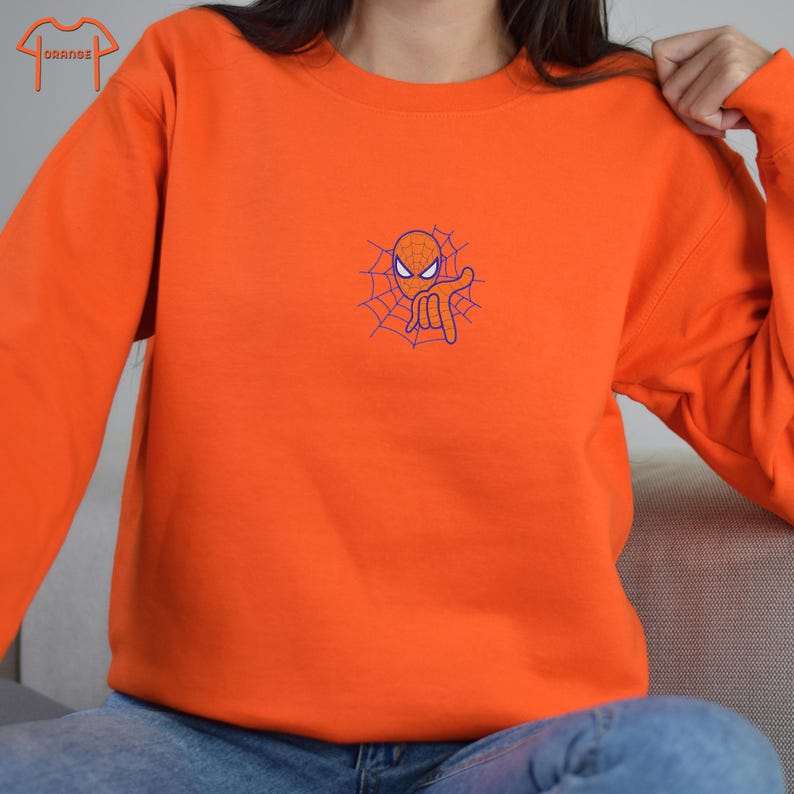 May include: Orange sweatshirt with a small embroidered image of Spiderman in a web. The image is in blue and orange.