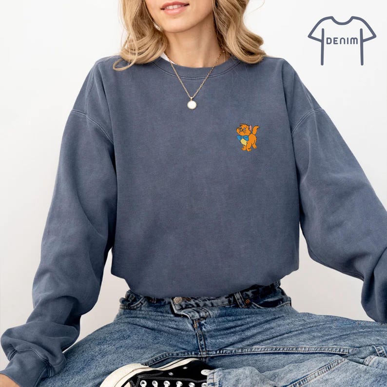 May include: A denim blue sweatshirt featuring a small cartoon dog graphic on the chest. The long-sleeved, crew-neck top is paired with blue jeans and black trainers. The word "DENIM" is printed on the top right.