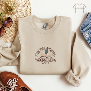 May include: A beige sweatshirt with the text "Flourish & Blotts Booksellers" in a brown, vintage-style font. The design includes an open book and a quill. The sweatshirt is made of a soft material.