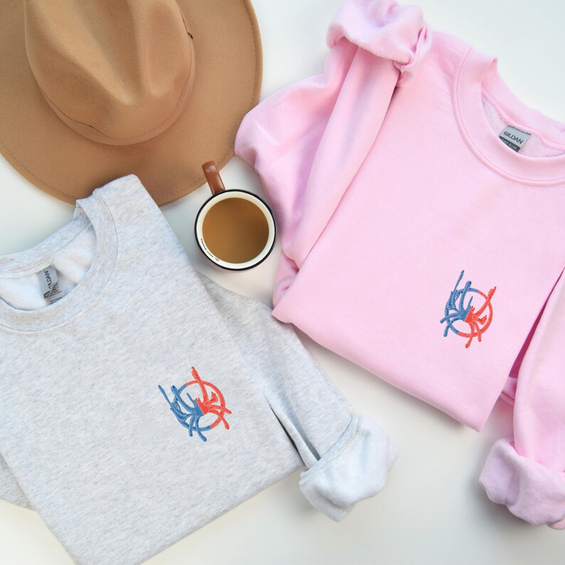 May include: Two sweatshirts featuring a graphic design of a red and blue circle with lines. One sweatshirt is grey and the other is pink.